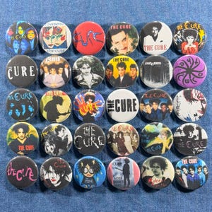 May include: A collection of 30 round pinback buttons featuring images of The Cure band members, album art, and the band's logo. The buttons are in various colors, including black, white, red, and blue, and are arranged on a denim surface.