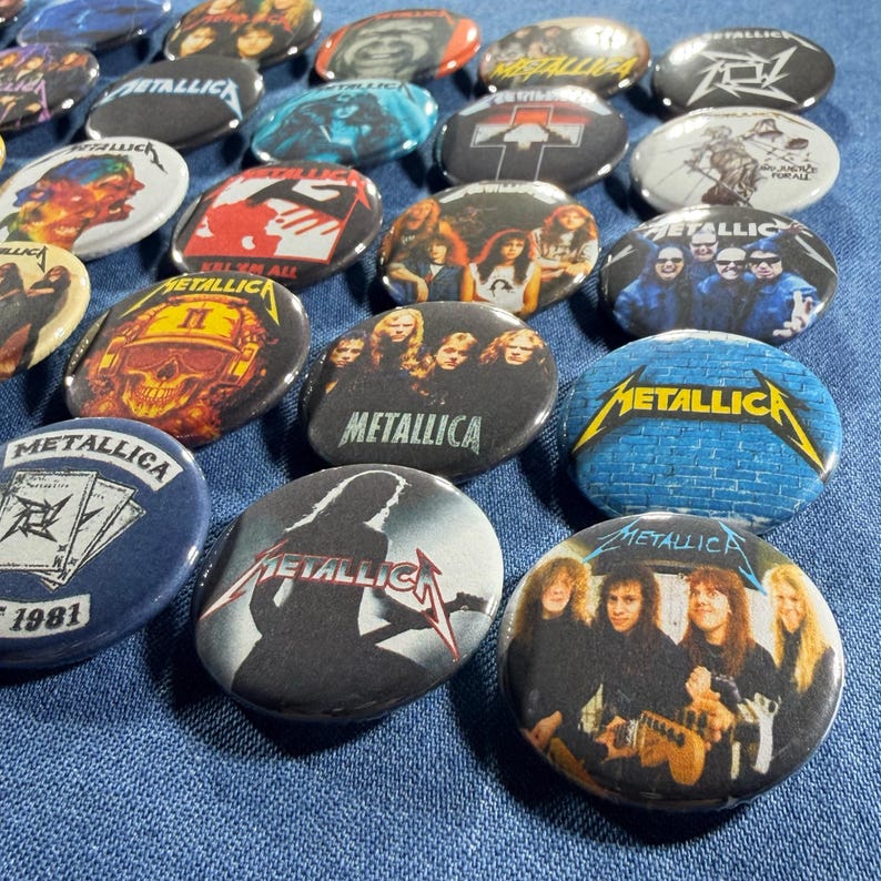 K&ouml;nnte beinhalten: Eine Sammlung runder Metall-Ansteckbuttons mit verschiedenen Designs und dem Bandnamen "Metallica". Die Buttons zeigen Bilder der Band, Albumcover und Logos in Farben wie Schwarz, Rot und Blau. Die Buttons sind auf einer blauen Stoffoberfl&auml;che angeordnet.
