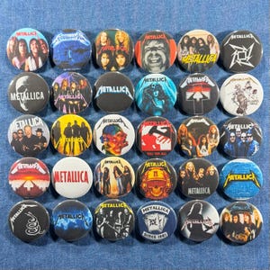 K&ouml;nnte beinhalten: Eine Sammlung runder Metallica-Bandbuttons mit verschiedenen Bildern und Logos. Die Buttons zeigen Bandmitglieder, Albumcover und das Metallica-Logo. Die Buttons befinden sich auf einem blauen Jeans-Hintergrund.