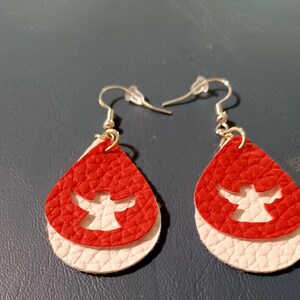 May include: A pair of dangle earrings with a red and white layered teardrop design. The teardrop is cut out with an angel shape.