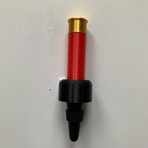 May include: A red and black shotgun shell bottle stopper. The top of the stopper is a gold-colored shotgun shell. The bottom of the stopper is a black plastic base with a pointed tip.