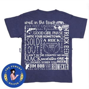 May include: A navy blue t-shirt with white text printed on the back. The text includes phrases like "wait in the truck", "six feet under", "good girl phase", "hate your hometown", "sold a rock out", "truck bed", "I ain't in the country no more", "jack rockstar one", "signed, sober you beer", "jim bob radio song", "rednecker", and "kill sh*t till I die".