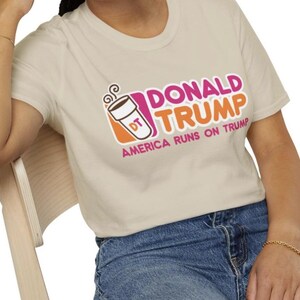 May include: A beige t-shirt with a pink and orange graphic of a coffee cup with the text "Donald Trump" and "America Runs on Trump".