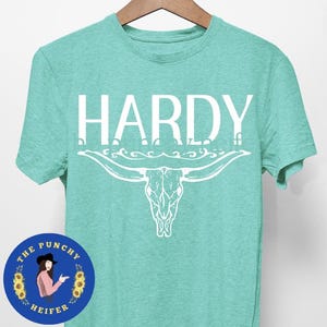 May include: A turquoise blue t-shirt with a white graphic of a bull skull with horns and the word "HARDY" above it. The t-shirt has a distressed look and is made of a soft, comfortable material.