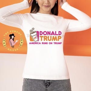 May include: White long-sleeved t-shirt with a pink and orange graphic of a coffee cup with the text "DONALD TRUMP" and "AMERICA RUNS ON TRUMP".