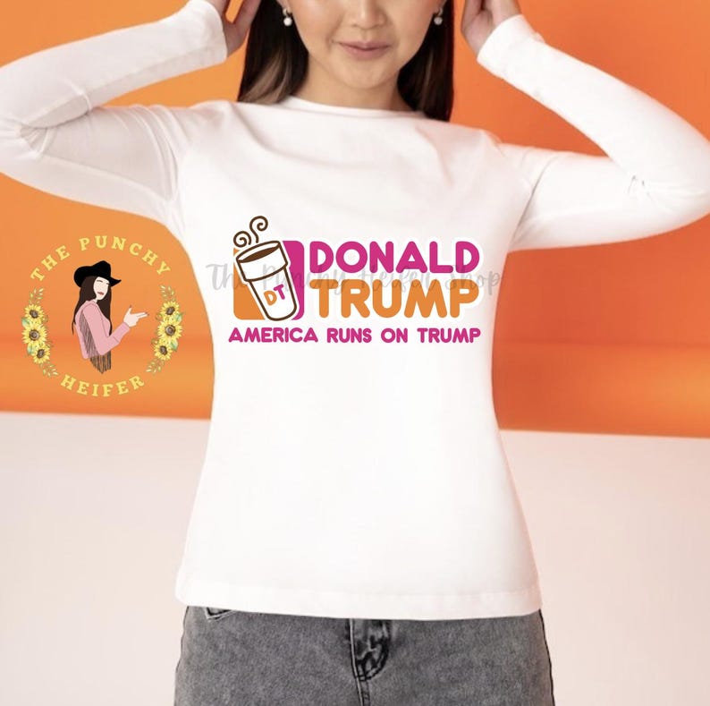 May include: White long-sleeved t-shirt with a pink and orange graphic of a coffee cup with the text "DONALD TRUMP" and "AMERICA RUNS ON TRUMP".