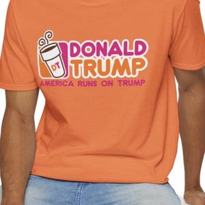 May include: Orange t-shirt with a pink and white graphic of a coffee cup with the text "Donald Trump" and "America Runs on Trump".