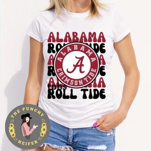 May include: White t-shirt with a retro design featuring the Alabama Crimson Tide logo and the text "Roll Tide".