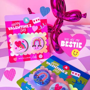 Może przedstawiać: Guziki i kartki z motywem walentynkowym. Na kartkach widnieje napis "Happy Valentine's Day" i "You are my Bestie". Guziki przedstawiają tęcze, serca i rolki. Schemat kolorów to różowy, fioletowy i niebieski.