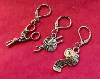 May include: Silver-toned earrings with lever-back closures. The charms are shaped like scissors, a ball of yarn with knitting needles, and a measuring tape. The earrings are displayed against a red background.