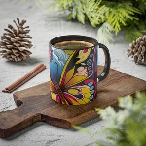 Peut inclure: Une tasse à café noire avec un motif de papillon coloré. Le papillon a des ailes jaunes, rouges, bleues et noires. La tasse est posée sur une planche à découper en bois.