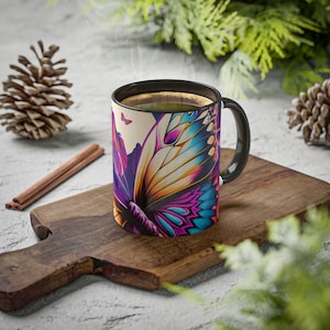 Könnte beinhalten: Eine Kaffeetasse mit schwarzem Henkel und einem lebendigen Schmetterlingsflügel-Design in Lila, Blau, Gelb und Pink. Die Tasse steht auf einem Holzbrett mit Zimtstangen und einem Tannenzapfen. Dampf steigt aus der dunklen Flüssigkeit im Inneren auf.