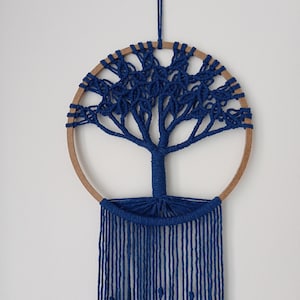 May include: A wall hanging featuring a circular wooden hoop with a dark blue macrame tree design inside. The tree trunk and branches are formed by knotted cord, with long, straight blue fringe hanging from the bottom of the hoop.