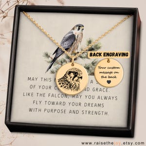 May include: Gold pendant necklace with a falcon illustration on a round charm. The necklace is displayed in a black box with printed text. The text reads "May this...fly toward your dreams." The necklace is presented in a gift box.