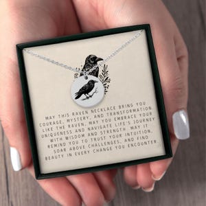 May include: A silver-toned circular pendant necklace featuring a black raven illustration. The pendant rests on a card printed with the text: MAY THIS RAVEN NECKLACE BRING YOU COURAGE, MYSTERY, AND TRANSFORMATION. LIKE THE RAVEN, MAY YOU EMBRACE YOUR UNIQUENESS.