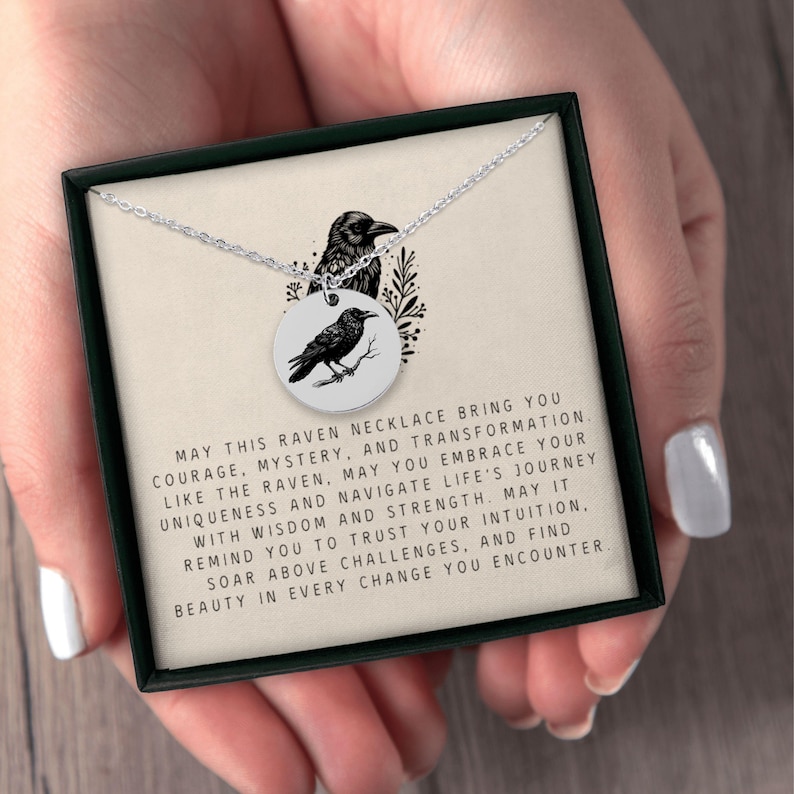 May include: A silver-toned circular pendant necklace featuring a black raven illustration. The pendant rests on a card printed with the text: MAY THIS RAVEN NECKLACE BRING YOU COURAGE, MYSTERY, AND TRANSFORMATION. LIKE THE RAVEN, MAY YOU EMBRACE YOUR UNIQUENESS.