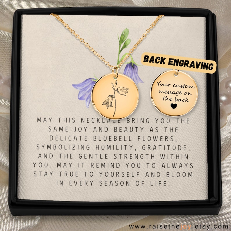 May include: A gold-toned circular pendant necklace featuring a delicate bluebell flower engraving on a chain. A second circular disc shows the text BACK ENGRAVING and Your custom message on the back with a heart icon. The card includes inspirational text.