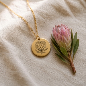 May include: A gold-toned necklace featuring a circular pendant engraved with a detailed illustration of a protea flower. The pendant hangs from a delicate chain, positioned next to a fresh pink and green protea flower bloom.