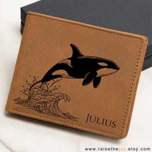 Peut inclure: Un portefeuille en cuir marron clair avec un motif d'orque noire sautant au-dessus des vagues. Le nom "JULIUS" est imprimé dans le coin inférieur droit. Le portefeuille est fermé et posé sur une surface sombre.
