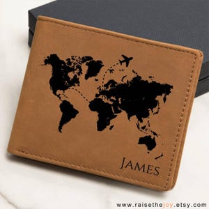 May include: A tan leather wallet with a black world map design and an airplane graphic. The name "JAMES" is printed in black on the lower right corner. The wallet is closed and sits on a white surface.