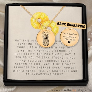 Op de afbeelding: Goudkleurige ketting met een ananas hanger, gepresenteerd in een zwarte doos. De hanger heeft een gedetailleerd ananas ontwerp. De doos bevat een kaart met inspirerende tekst. Een tweede hanger is afgebeeld met de tekst "Back Engraving".
