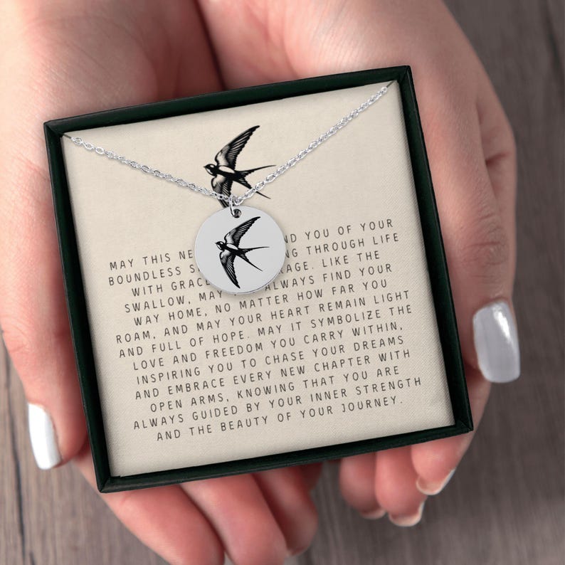 May include: A silver necklace with a round pendant featuring a black and white silhouette of a swallow. The pendant is on a white card with an inspirational message about finding your way home and chasing your dreams. The message reads: "May this necklace be a boundless source of grace with you. May the swallow, may it always find your way home. No matter how far you roam, and may your heart remain light and full of hope. May it symbolize the love and freedom you carry within, inspiring you to chase your dreams and embrace every new chapter with open arms, knowing that you are always guided by your inner strength and the beauty of your journey."