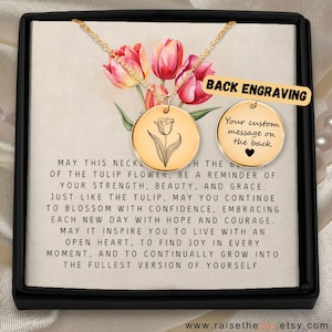 Può includere: Una collana color oro con un ciondolo rotondo con un'illustrazione di tulipano. Il ciondolo è esposto in una scatola nera con una carta che riporta un testo sul fiore del tulipano. Sono visibili anche le parole "BACK ENGRAVING".