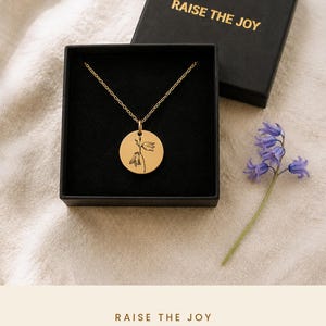 May include: A gold-toned circular pendant necklace featuring a delicate black line-art engraving of a bluebell flower. The necklace is presented in a black square gift box with the text RAISE THE JOY printed in gold on the lid.