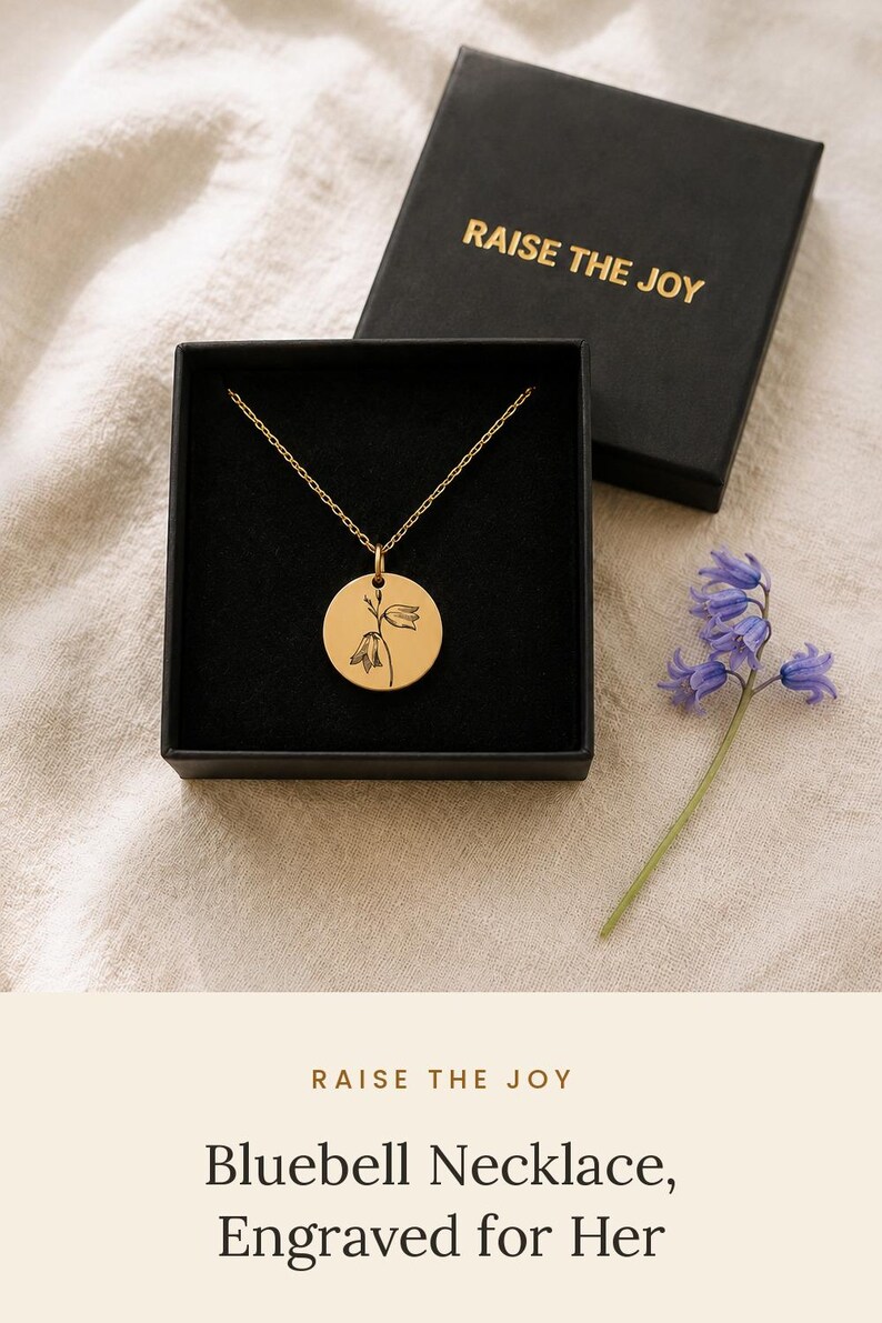 May include: A gold-toned circular pendant necklace featuring a delicate black line-art engraving of a bluebell flower. The necklace is presented in a black square gift box with the text RAISE THE JOY printed in gold on the lid.