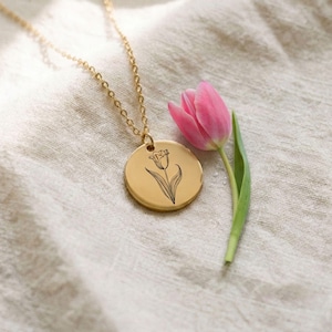 Könnte beinhalten: Eine goldfarbene Halskette mit rundem Anhänger, der eine feine schwarze Strichzeichnung einer Tulpe zeigt. Der Anhänger hängt an einer passenden dünnen Goldkette und liegt neben einer einzelnen frischen rosa Tulpe mit grünem Stiel.