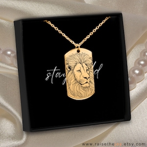 Könnte beinhalten: Goldfarbene Halskette mit Hundemarkenanhänger und detaillierter Löwenkopf-Gravur. Die Halskette wird in einer schwarzen Box mit dem Schriftzug "stay wild" in Schreibschrift präsentiert. Der Hintergrund ist ein Satin-Stoff.