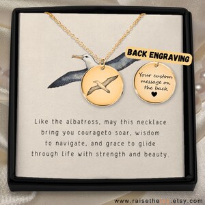 Op de afbeelding: Goudkleurige ketting met een ronde hanger met een gegraveerde albatros. De ketting wordt gepresenteerd in een zwarte doos met de tekst: "Zoals de albatros, moge deze ketting je de moed geven om te stijgen..." De hanger is ongeveer 2,5 cm in diameter.