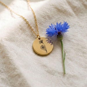 Könnte beinhalten: Eine goldfarbene Halskette mit rundem Anhänger, der eine feine schwarze Strichzeichnung einer Wildblume zeigt. Der Anhänger hängt an einer passenden goldfarbenen Kette und liegt neben einer einzelnen, leuchtend blauen Kornblume.