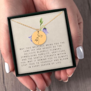 May include: A gold-toned circular pendant necklace featuring a black line drawing of a bluebell flower. The pendant rests on a card printed with text about bluebell flowers, humility, gratitude, and inner strength, all inside a black box.