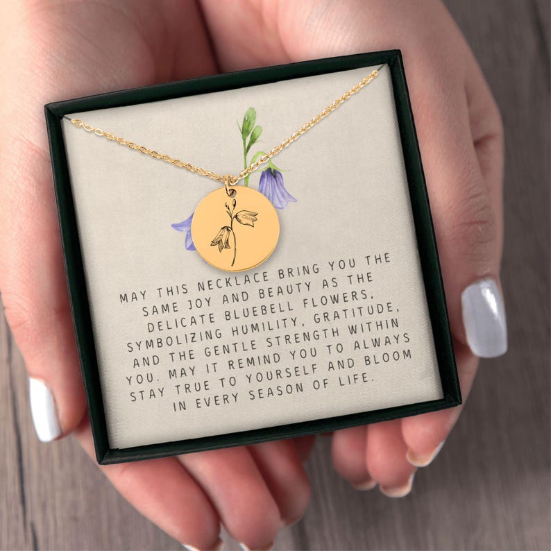 May include: A gold-toned circular pendant necklace featuring a black line drawing of a bluebell flower. The pendant rests on a card printed with text about bluebell flowers, humility, gratitude, and inner strength, all inside a black box.