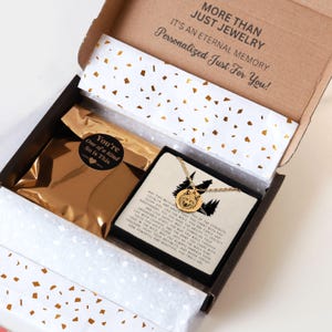 K&ouml;nnte beinhalten: Ge&ouml;ffnete Geschenkbox mit einer goldenen Halskette mit Wolf-Anh&auml;nger. Die Box enth&auml;lt einen goldenen Beutel, eine Karte mit Text und Seidenpapier mit goldenem Konfetti. Der Text auf der Box lautet: "MORE THAN JUST JEWELRY".