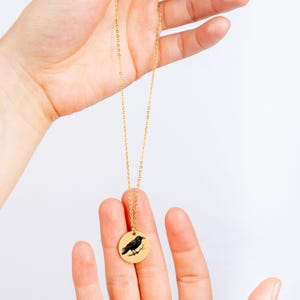 May include: A gold-toned necklace featuring a thin chain and a circular pendant. The pendant displays a black engraved illustration of a crow perched on a branch. The metal surface of the pendant is smooth and reflective, contrasting with the dark crow.