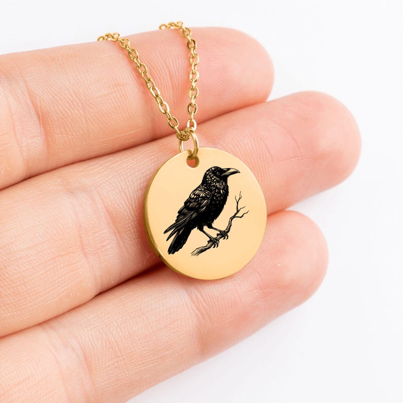 May include: A gold-toned circular pendant necklace featuring a detailed black engraved illustration of a raven perched on a thin branch. The pendant hangs from a matching gold-toned metal chain, showcasing a clean and polished metallic finish.