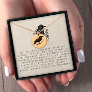 May include: A gold-toned circular pendant necklace featuring a black raven illustration, presented in a black gift box with a printed card that reads: MAY THIS RAVEN NECKLACE BRING YOU COURAGE, MYSTERY, AND TRANSFORMATION. LIKE THE RAVEN, MAY YOU EMBRACE YOUR UNIQUENESS AND NAVIGATE LIFE'S JOURNEY WITH WISDOM AND STRENGTH. MAY IT REMIND YOU TO TRUST YOUR INTUITION, SOAR ABOVE CHALLENGES, AND FIND BEAUTY IN EVERY CHANGE YOU ENCOUNTER.