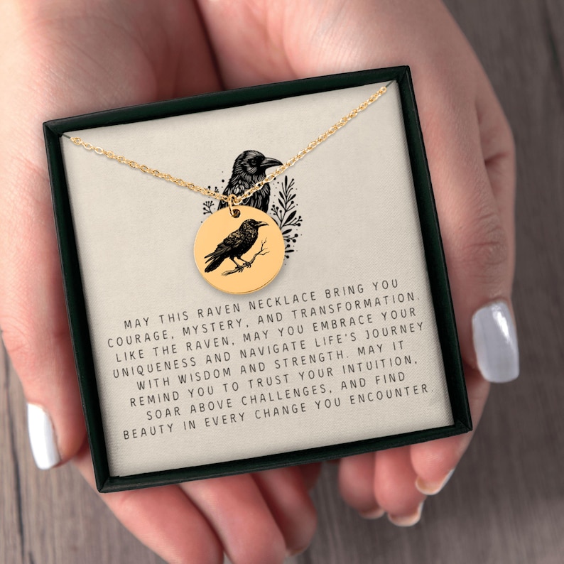 May include: A gold-toned circular pendant necklace featuring a black raven illustration, presented in a black gift box with a printed card that reads: MAY THIS RAVEN NECKLACE BRING YOU COURAGE, MYSTERY, AND TRANSFORMATION. LIKE THE RAVEN, MAY YOU EMBRACE YOUR UNIQUENESS AND NAVIGATE LIFE'S JOURNEY WITH WISDOM AND STRENGTH. MAY IT REMIND YOU TO TRUST YOUR INTUITION, SOAR ABOVE CHALLENGES, AND FIND BEAUTY IN EVERY CHANGE YOU ENCOUNTER.