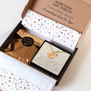 May include: A gold-toned necklace with a circular pendant featuring a bluebell flower engraving, presented on a card with text inside a cardboard gift box. The box includes a gold metallic pouch with a sticker reading You're One of a Kind.