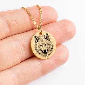K&ouml;nnte beinhalten: Goldfarbene Halskette mit Anh&auml;nger, die eine detaillierte Schwarz-Wei&szlig;-Illustration eines Wolfskopfes zeigt. Der runde Anh&auml;nger h&auml;ngt an einer zarten Goldkette. Das Fell des Wolfes ist aufwendig gezeichnet, und der Anh&auml;nger hat eine polierte Oberfl&auml;che.