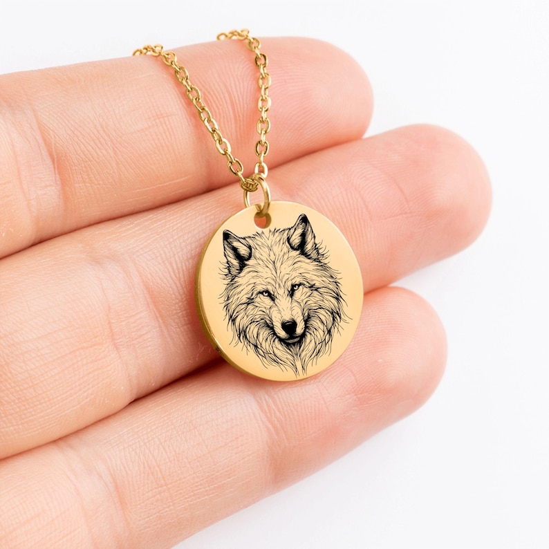 K&ouml;nnte beinhalten: Goldfarbene Halskette mit Anh&auml;nger, die eine detaillierte Schwarz-Wei&szlig;-Illustration eines Wolfskopfes zeigt. Der runde Anh&auml;nger h&auml;ngt an einer zarten Goldkette. Das Fell des Wolfes ist aufwendig gezeichnet, und der Anh&auml;nger hat eine polierte Oberfl&auml;che.