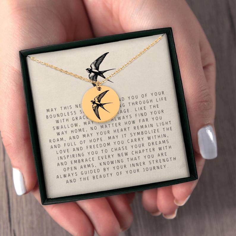 May include: A gold necklace with a pendant featuring a black and white silhouette of a bird. The pendant is in a box with a message that reads: "May this new boundless strength with grace swallow, may you find your way home, no matter how far you roam, and may your heart remain light and full of hope. May it symbolize the love and freedom you carry within, inspiring you to chase your dreams and embrace every new chapter with open arms, knowing that you are always guided by your inner strength and the beauty of your journey."