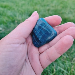 May include: A teal blue gemstone with a striated pattern, held in a hand against a green grassy background.