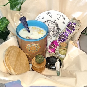 May include: A gift box filled with various items for spiritual practices, including a candle, a crystal pendulum, a smudge stick, a crystal, a small bottle of herbs, and a wooden coaster. The box is decorated with a light purple and white pattern.