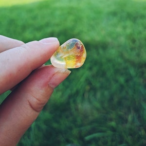 Puede incluir: Una gema transparente y translúcida con inclusiones amarillas y naranjas. La gema se sostiene en una mano contra un fondo de hierba verde.