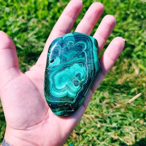 Puede incluir: Una piedra de malaquita lisa y verde con patrones en espiral. La piedra es una pieza natural, pulida y de forma ovalada.