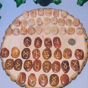 Può includere: Un set di 25 pietre di corniola arancione con rune dorate incise su ciascuna. Le pietre sono disposte in cerchio su una superficie di legno.