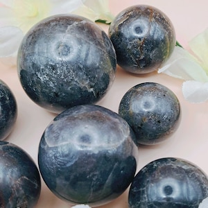 Peut inclure: Plusieurs sphères gris foncé avec des motifs uniques et des variations de couleur. Les sphères sont de tailles différentes et sont disposées sur une surface rose pâle, avec des décorations florales blanches.